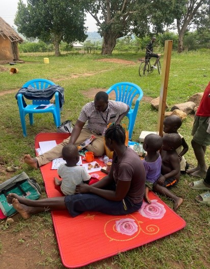 Children and two adults sit on a red mat outdoors, engaging in activities as part of a parenting program in Tororo.